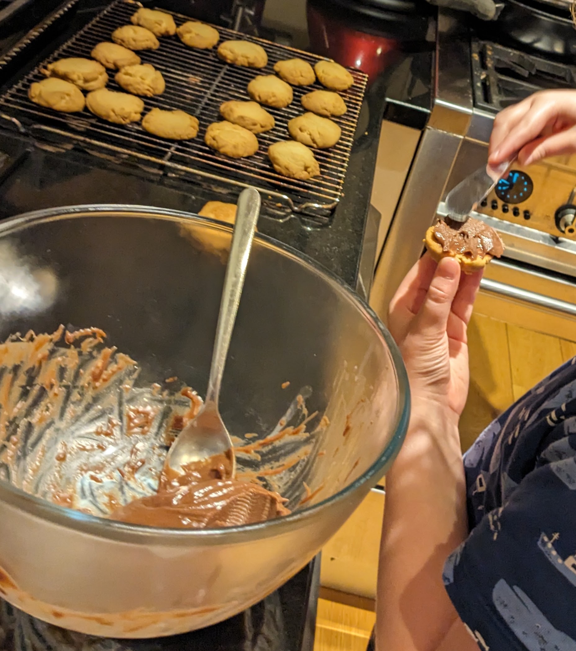 Picture of a tray of biscuit halves, with one getting the chocolate filling carefully added from the bowl of chocolate ganache.