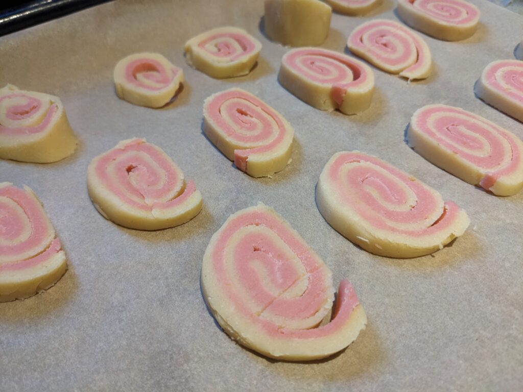 Slices of dough showing alternate swirls of pink and white on baking paper on a tray ready for the oven.